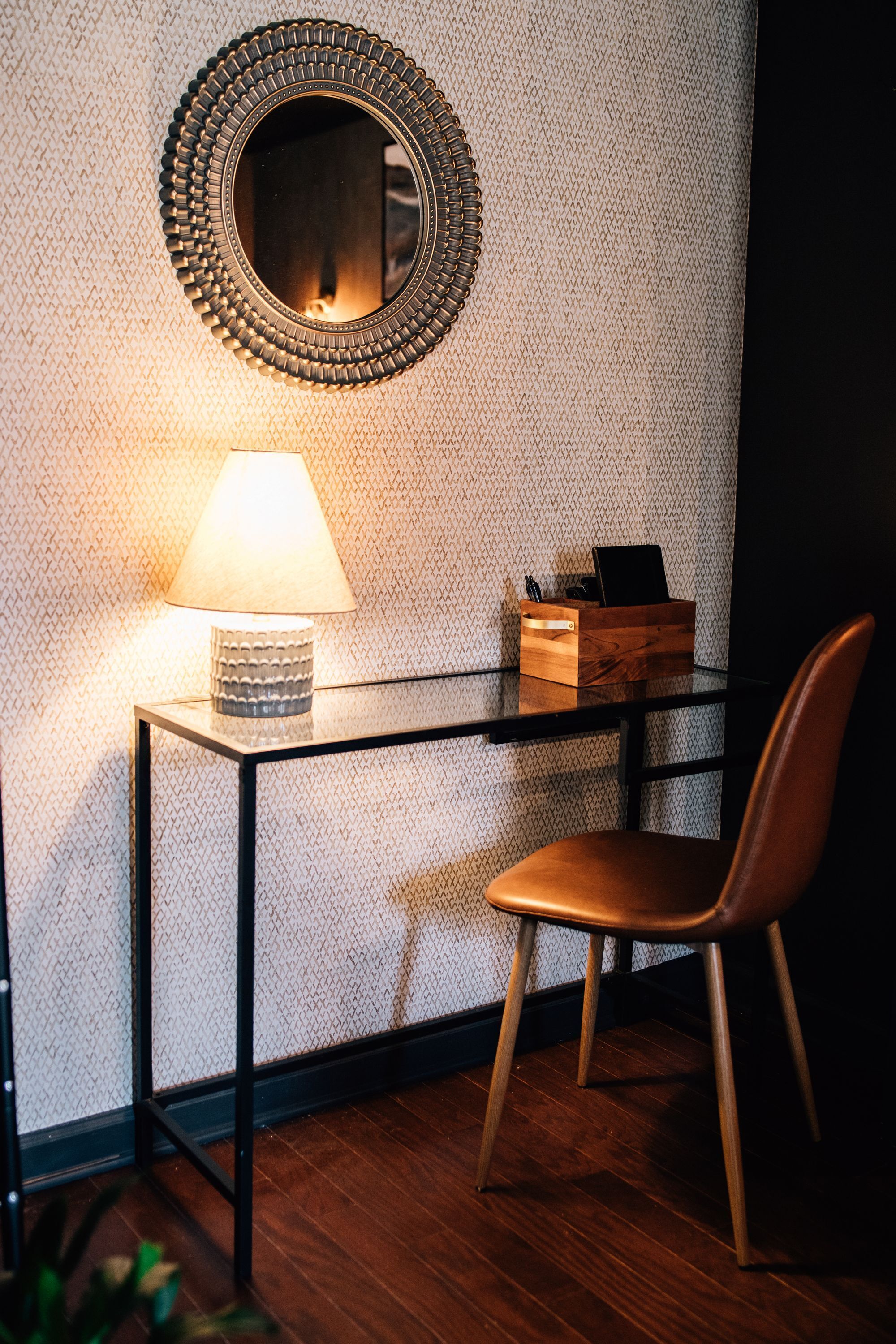 A small, modern room with a round mirror, a table with a lamp, and a wooden chair.