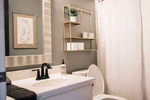 A bathroom with a white sink, black faucet, and a toilet. There is a framed picture on the wall and a shelf with toiletries.
