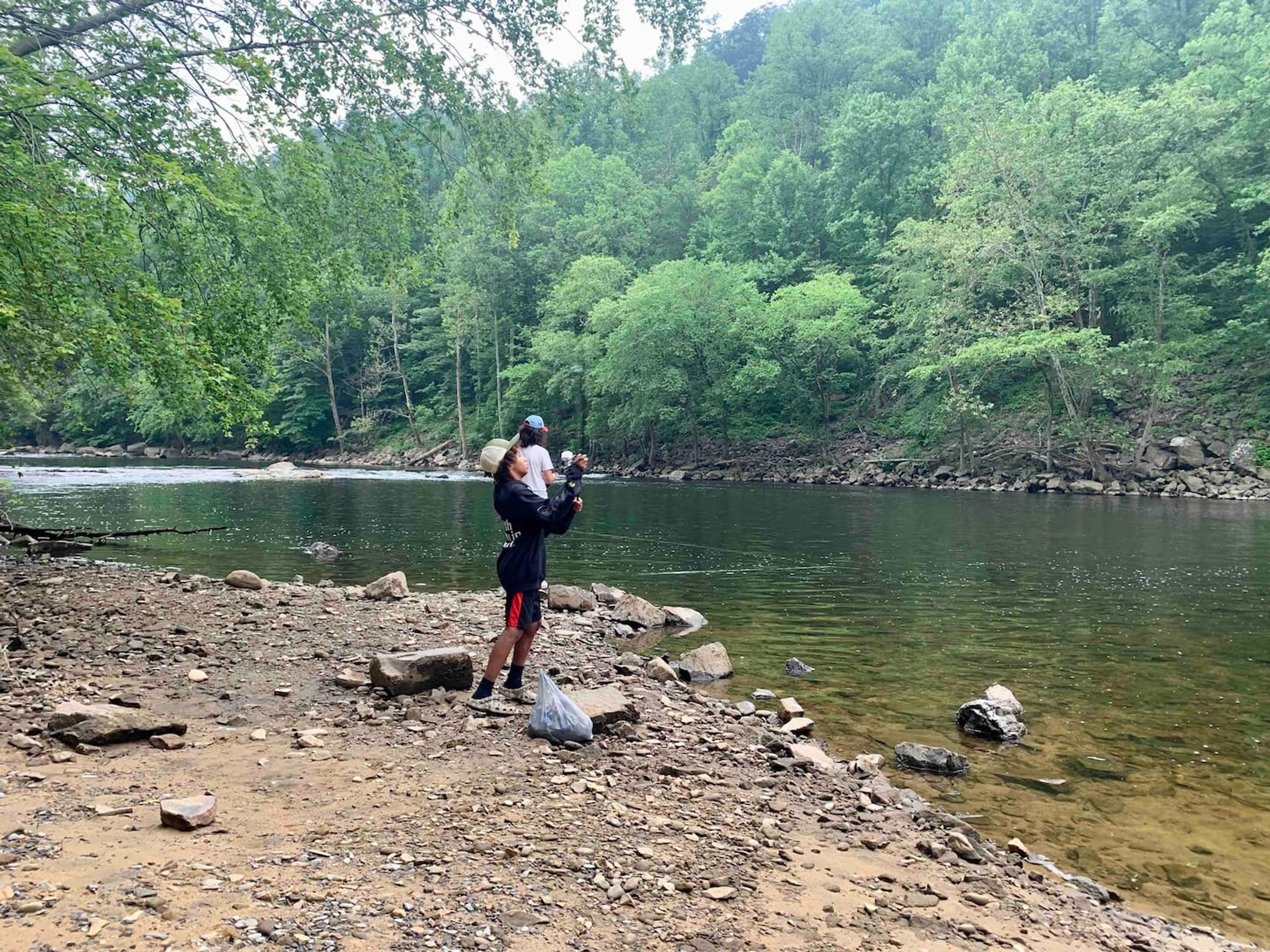 A rocky riverbank with a person standing by the water, surrounded by dense green trees.