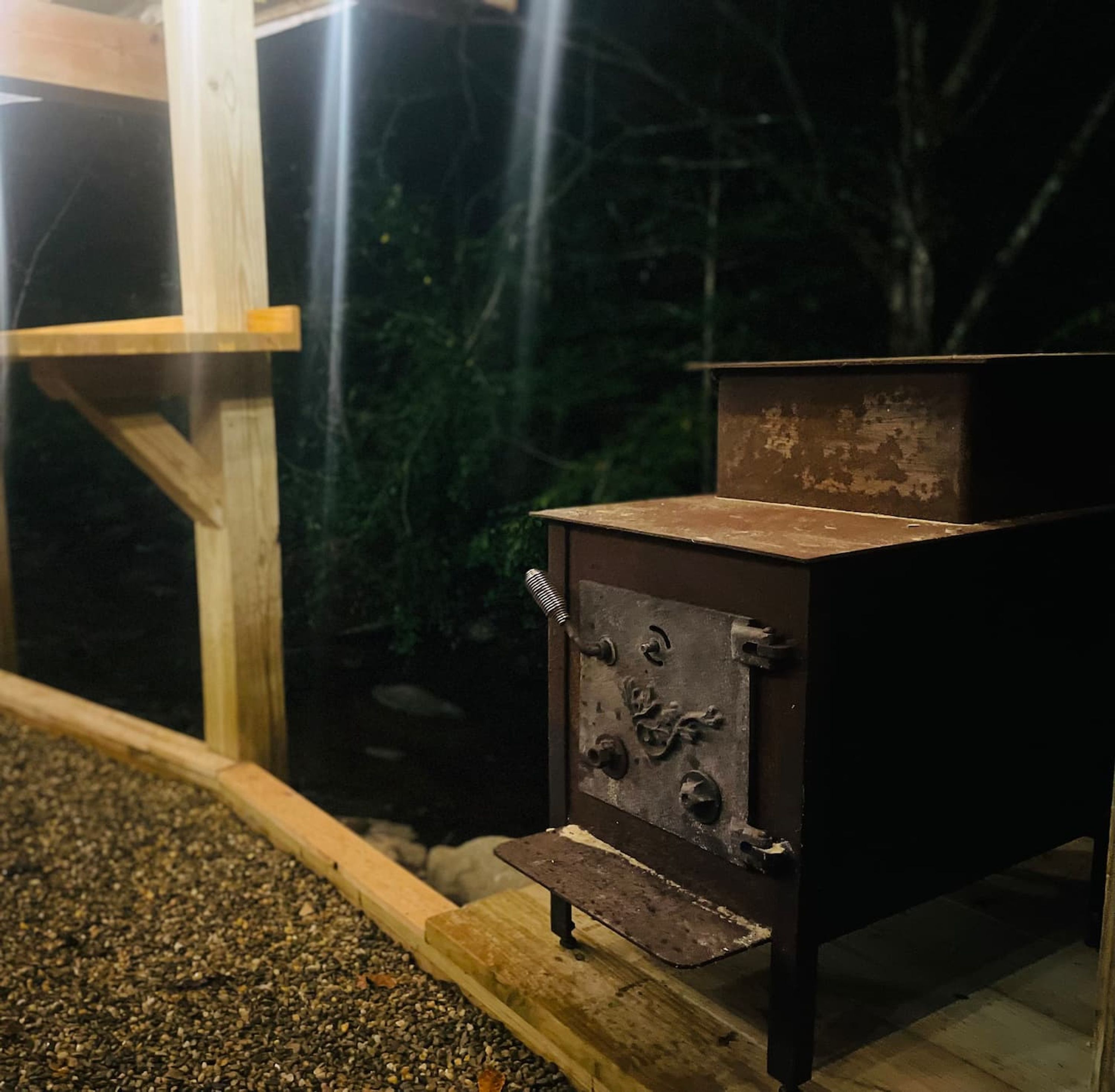 A small, rustic outdoor cooking stove with a metal grate, set against a backdrop of trees and a wooden structure.