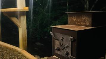 A small, rustic outdoor cooking stove with a metal grate, set against a backdrop of trees and a wooden structure.