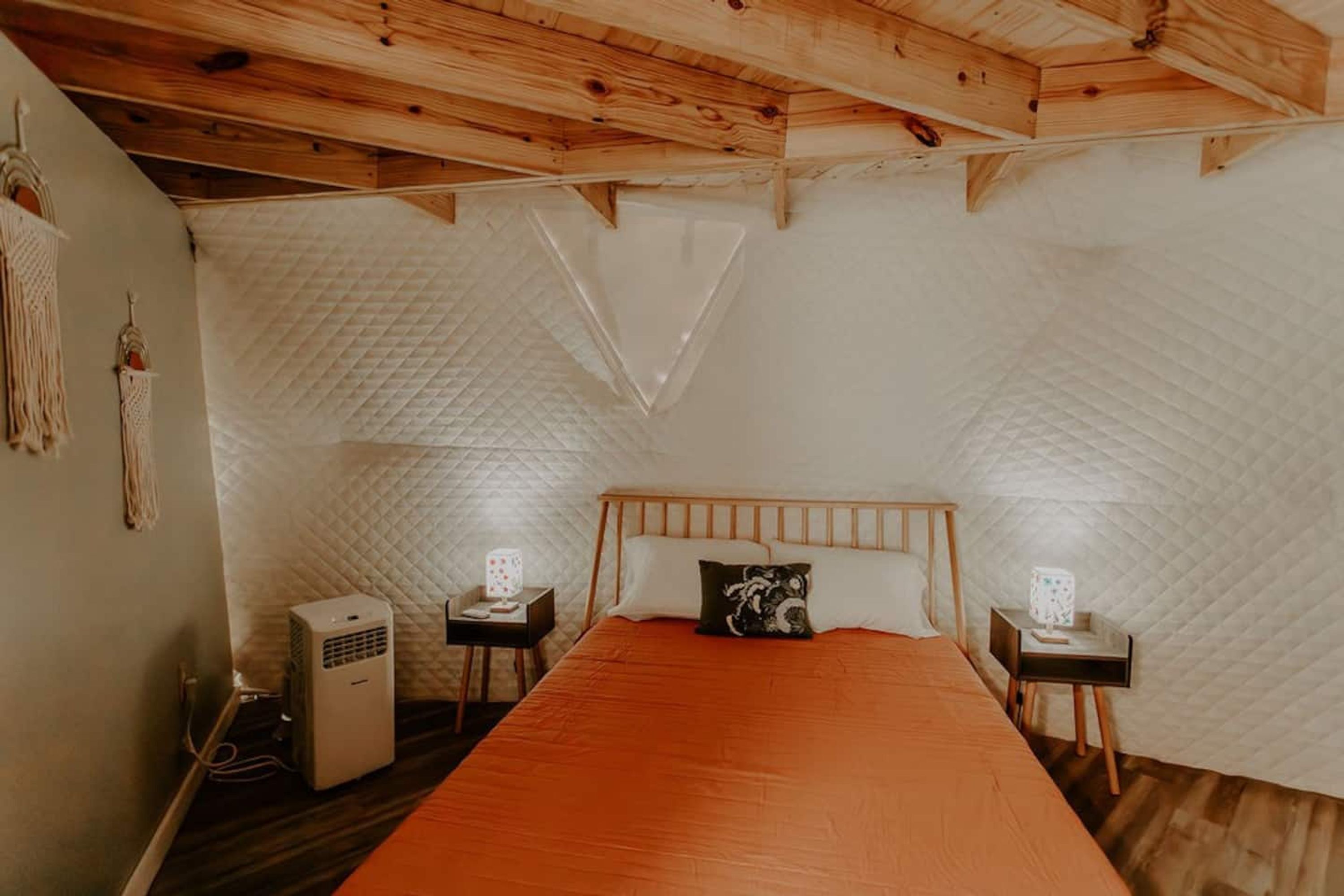 Un dormitorio con techo de madera, pared acolchada, cama con cubrecama naranja, dos mesitas de noche con lámparas y un equipo de aire acondicionado.