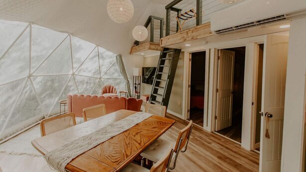 A cozy interior with a wooden dining table, cushioned chairs, a ladder leading to a loft, and a large window with a geometric view. - Slate Crossing Creekside Dome (Cosby)