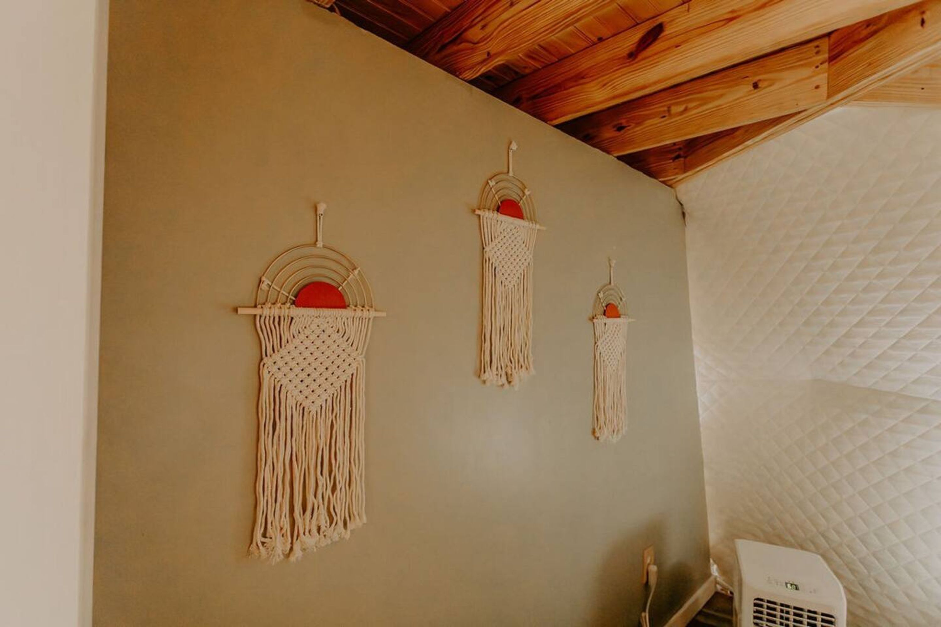 A room with a wooden ceiling, three macramé wall hangings, and a white quilted bed.