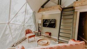 A spacious living area with a large window, a sofa, a coffee table, and a television mounted on the wall. - Slate Crossing Creekside Dome (Cosby)