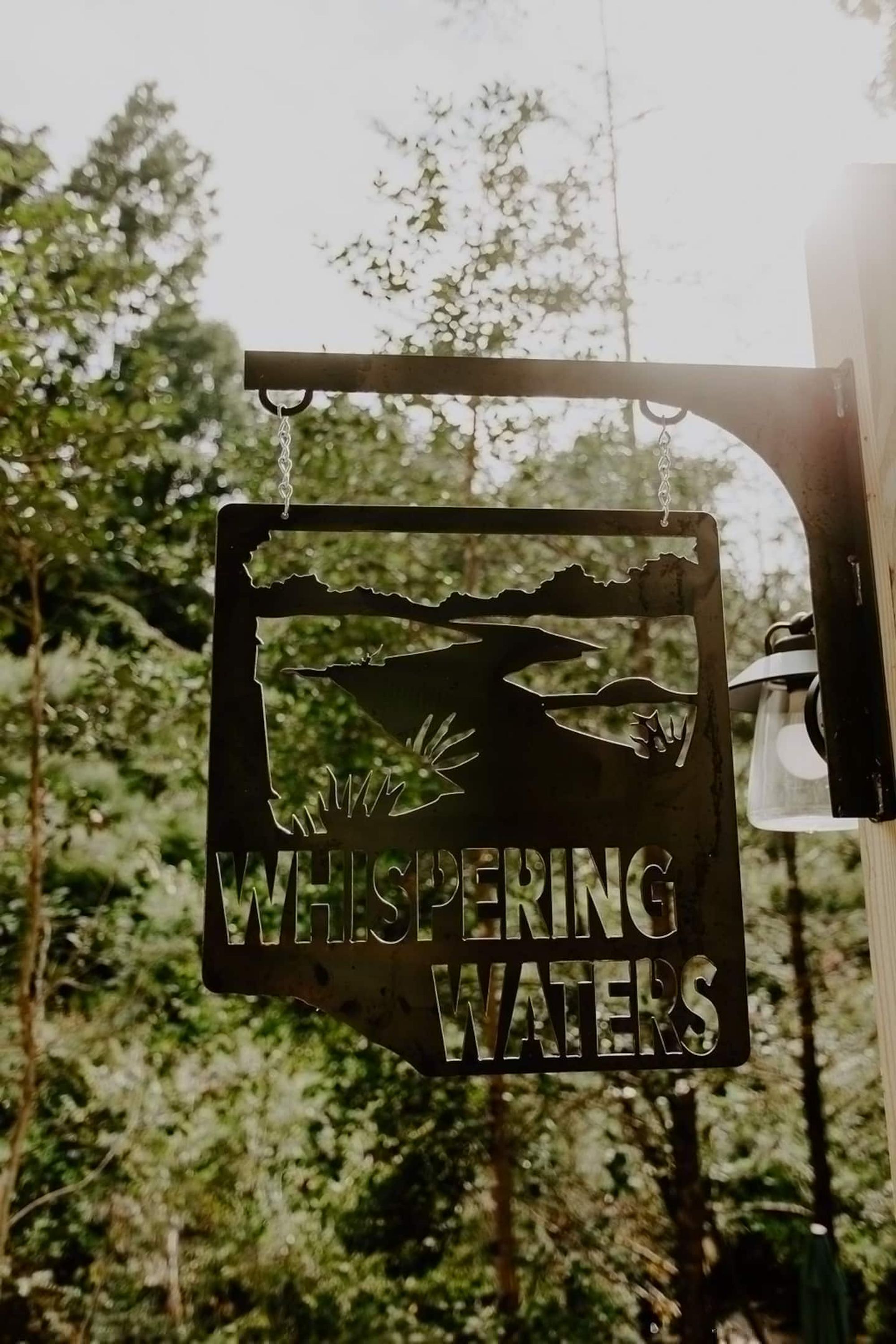 A metal sign with the text "WHISPERING WATERS" and a silhouette of a landscape.