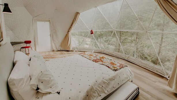 A cozy bedroom with a large window, a bed with a floral bedspread, a red chair, and a lamp. - Whispering Waters Dome Getaway (Cosby)