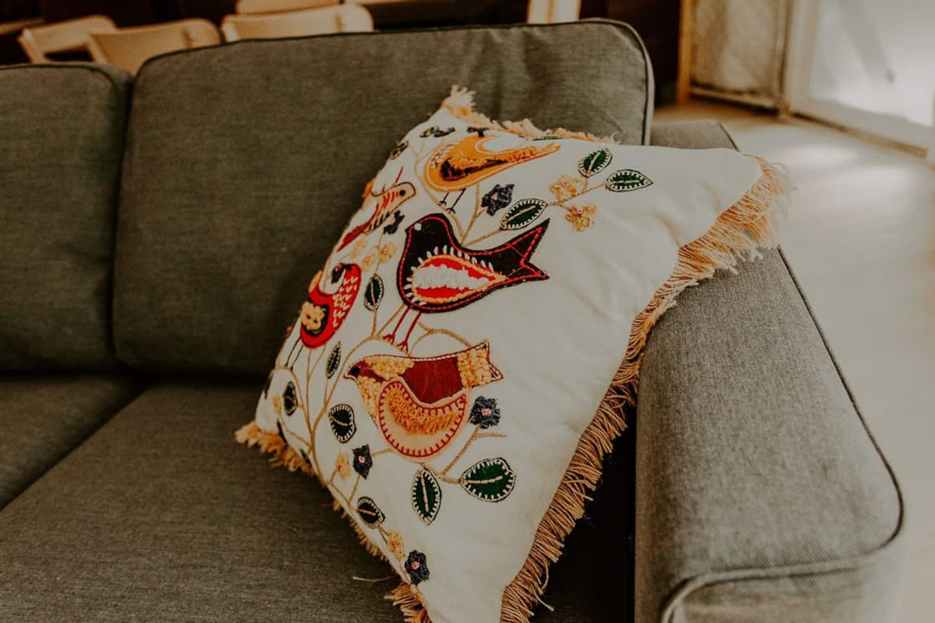 A grey sofa with a white cushion featuring embroidered birds and leaves.