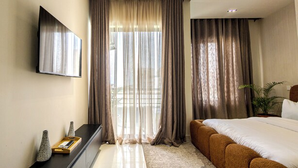 Une chambre à coucher moderne, dotée d’un grand lit, d’un téléviseur à écran plat fixé au mur et d’une grande fenêtre agrémentée de rideaux légers.