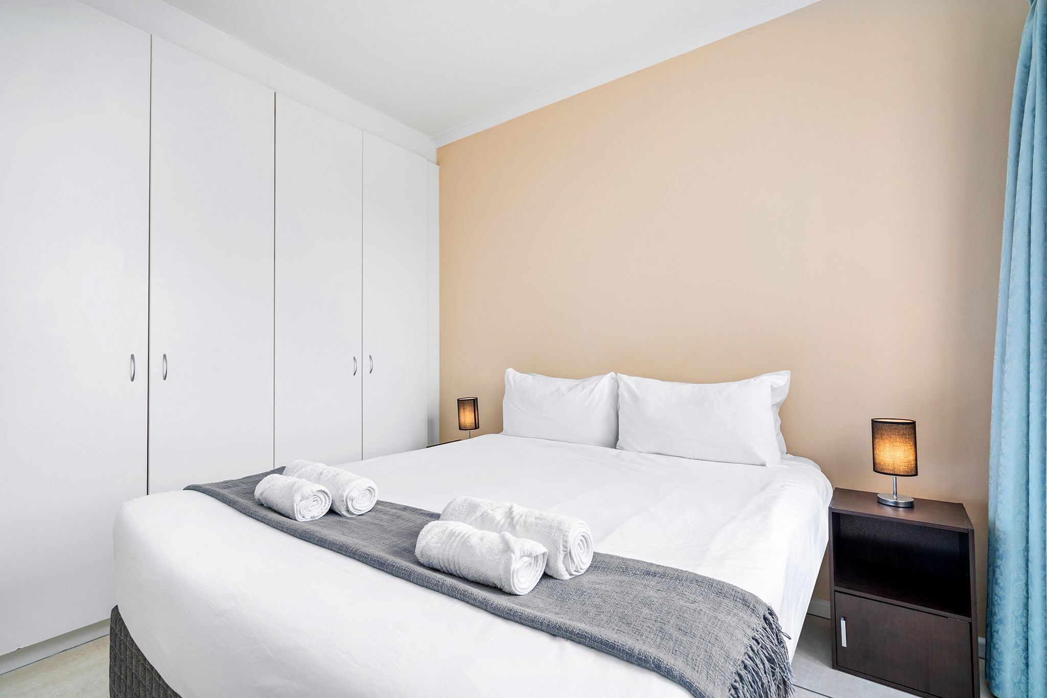 Hotelkamer met een groot bed, witte lakens, een grijze plaid, twee opgerolde handdoeken, een nachtkastje met een lamp en een kledingkast.