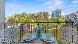Un balcón con dos sillas blancas, una mesa con un florero y vistas a palmeras y edificios.