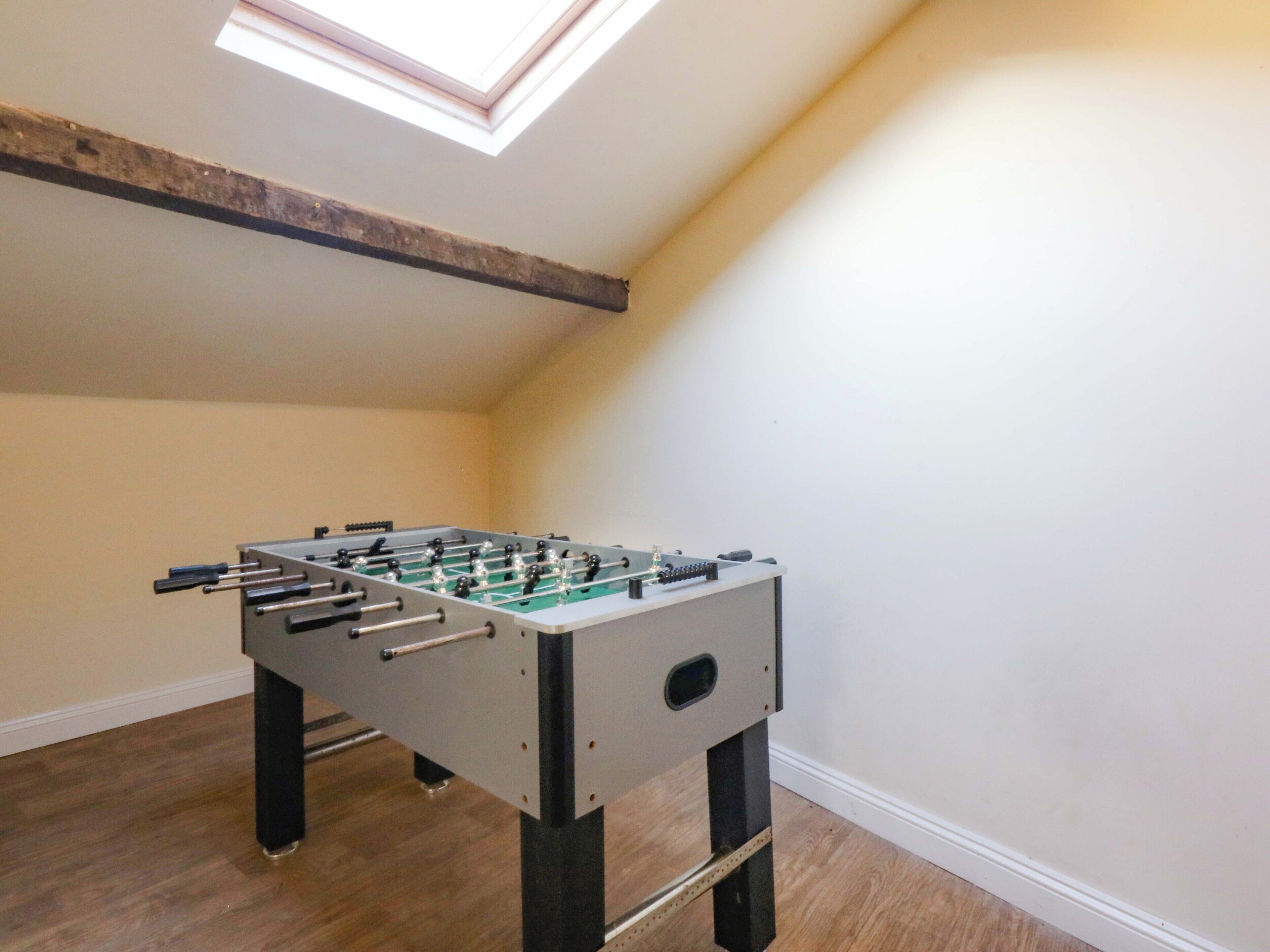 A room with a foosball table, wooden flooring, a sloped ceiling with a skylight, and a white wall.