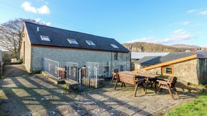 Ein Steinhaus mit geneigtem Dach und Dachfenstern, ein umzäunter Patio-Bereich mit Holztisch und Bänken sowie eine Bergblick im Hintergrund.