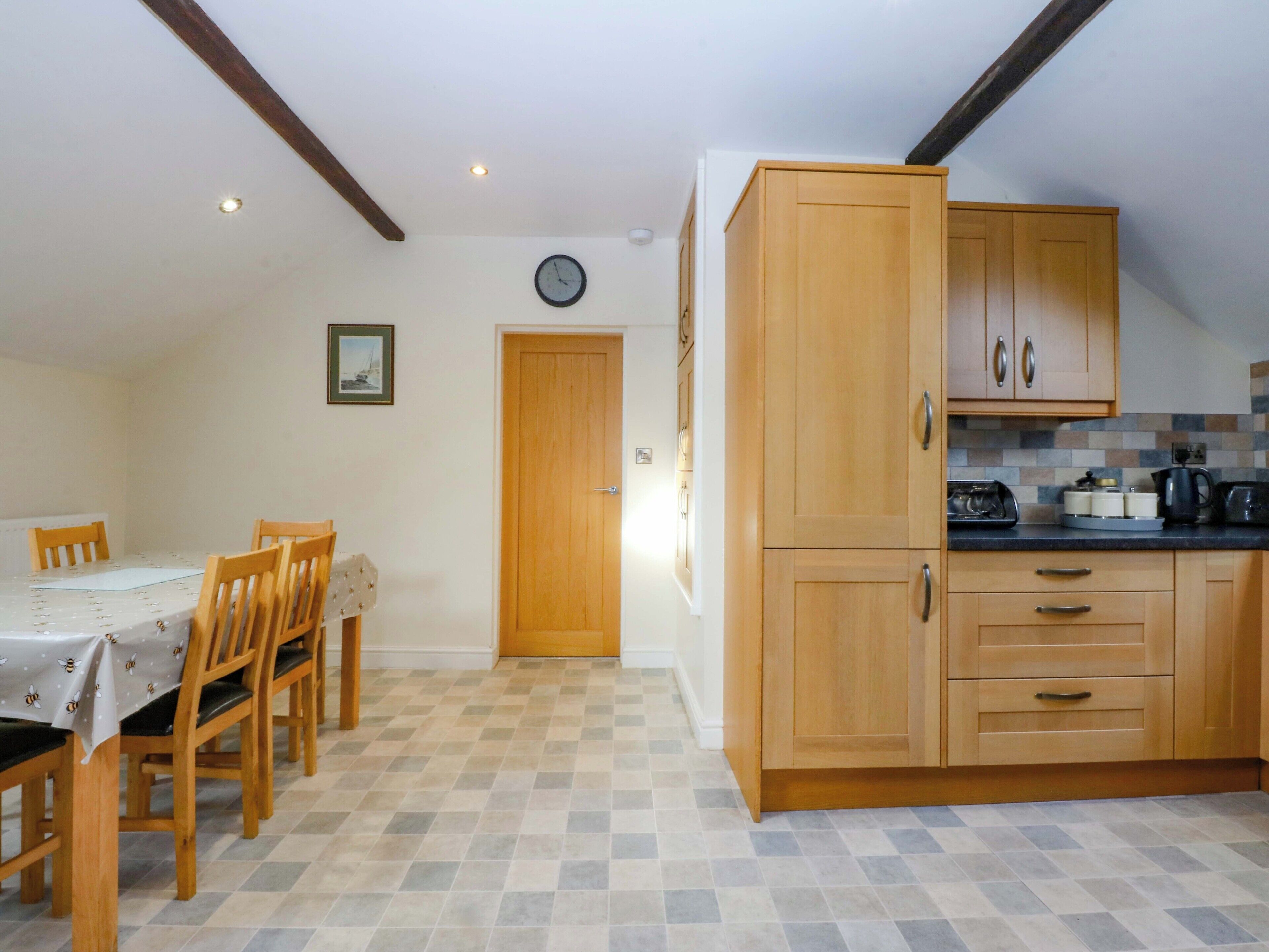 A dining area with a table and chairs, a kitchen with wooden cabinets and a tiled backsplash, and a clock on the wall.