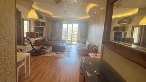 A spacious living area with a wooden floor, a sofa set, a coffee table, and a TV. There is a large mirror on the wall and a staircase leading to another level.