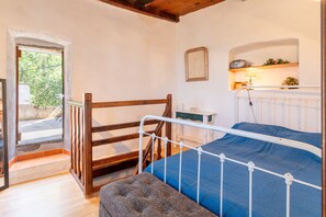 A bedroom with a bed, a wooden bench, a white radiator, a small table with a lamp, and a window with a view of greenery.