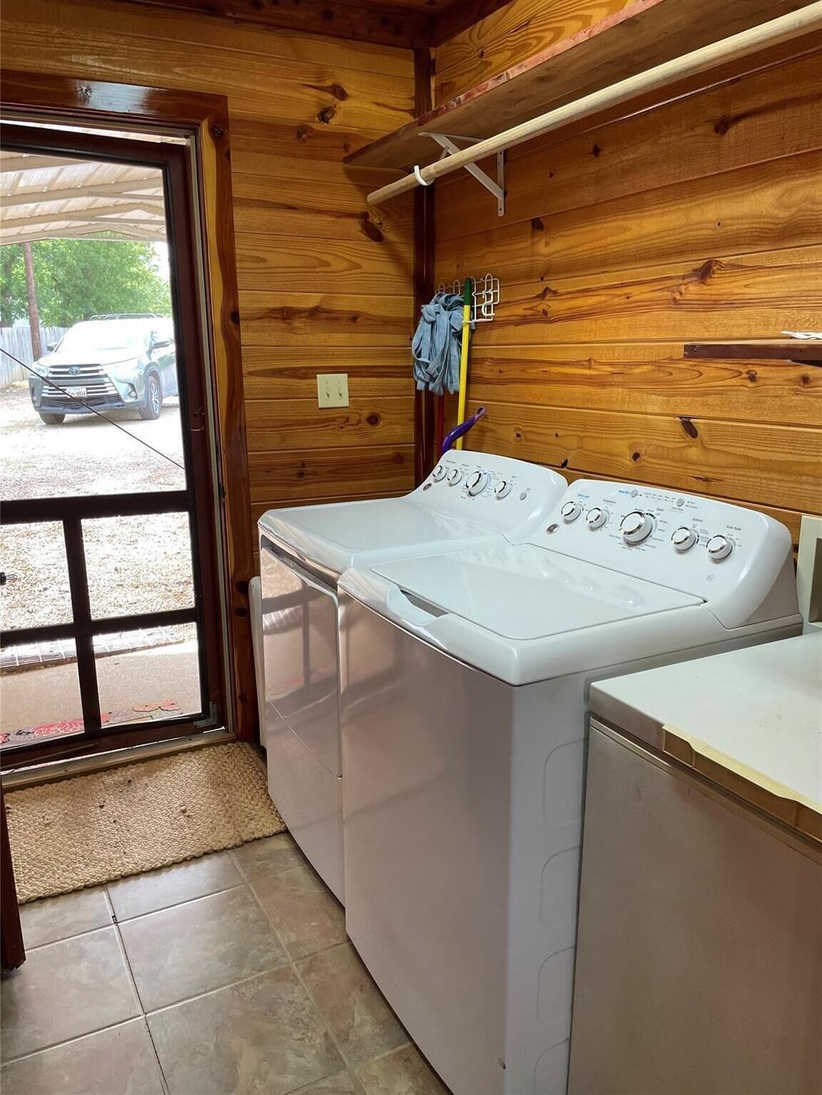 A laundry room with a front-loading washer and dryer, wooden walls, a window with a view of a parked car outside, and a hanging rack above the dryer.