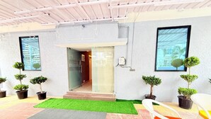A hotel entrance with a glass door, potted plants, a green artificial turf, and a white chair with an orange cushion. - Rasa Boutique Stay (Greater Noida)