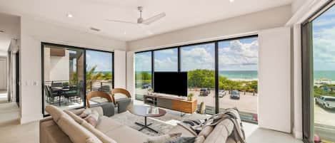 A modern living room with a large TV, comfortable seating, and a view of the beach.