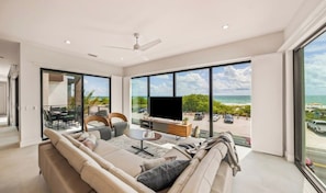 A modern living room with a large TV, comfortable seating, and a view of the beach.