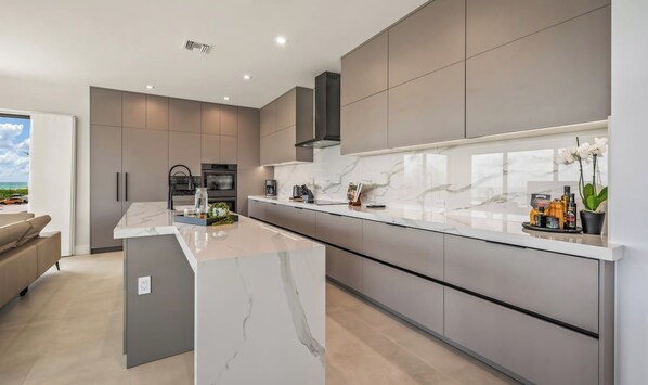 A modern kitchen with a marble island, grey cabinetry, and a view of the ocean.