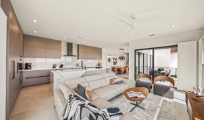 Eine moderne Küche mit einer zentralen Kochinsel, ein Essbereich mit einer Couch und Sesseln, sowie ein Balkon mit Sitzgelegenheit im Freien.