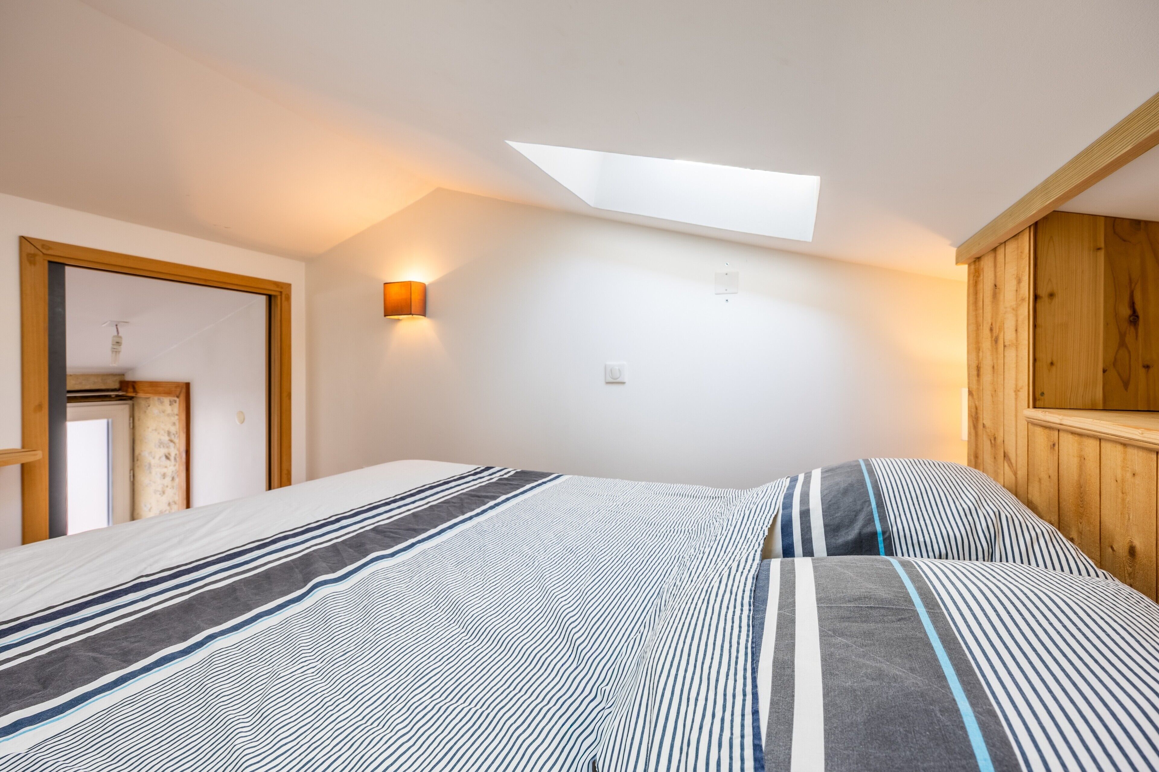 Une chambre avec un puits de lumière, un lit avec une literie à rayures, une armoire en bois et un luminaire fixé au mur.
