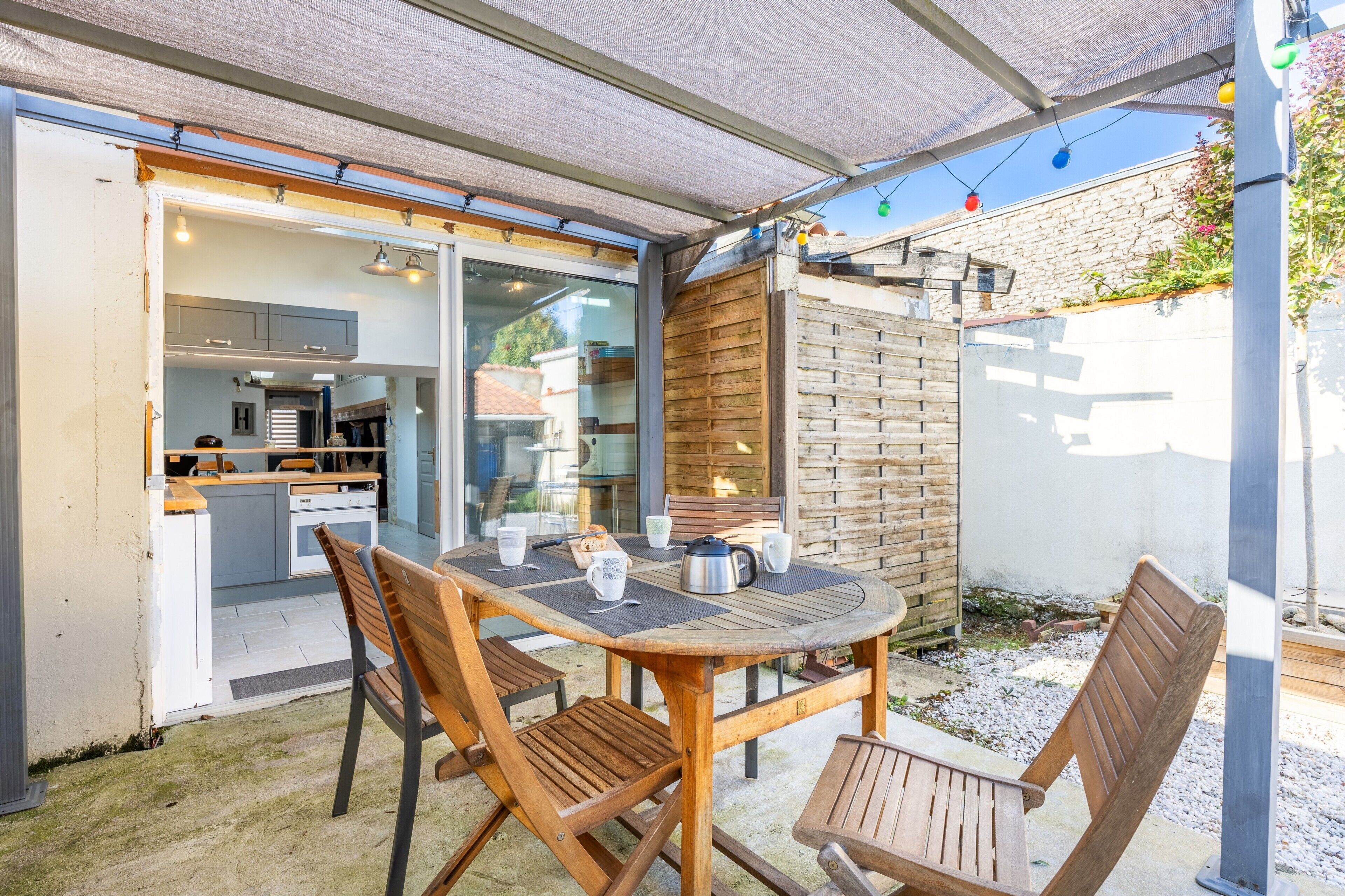 Une terrasse extérieure avec une table et des chaises, une clôture en bois et une vue sur l’intérieur de la maison.