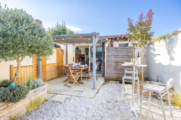 A patio with a wooden table and chairs, a pergola with string lights, and a small garden area with a tree and shrubs.
