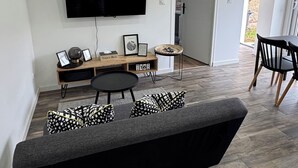 A modern living room with a large flat-screen TV mounted on the wall, a dark grey sofa with patterned cushions, a small wooden side table, and a dining area with a table and chairs. - Getaway 2 steps from the Loire - Loft Ligérien (Sandillon)