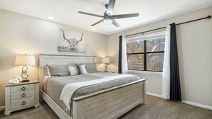 Un dormitorio con una cama de madera, dos mesitas de noche con lámparas, un ventilador de techo, una ventana con cortinas y una obra de arte colgada en la pared.
