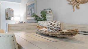 A dining table with a woven basket centerpiece, a white cushioned bench, a framed artwork, and a white birdcage with plants. - Oceanwalk 9-302 (New Smyrna Beach)