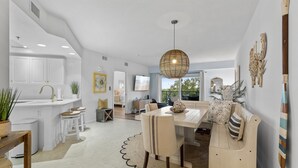 A modern kitchen with white cabinets, a dining area with a wooden table and chairs, and a large window providing natural light. - Oceanwalk 9-302 (New Smyrna Beach)