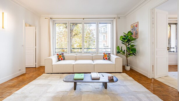 A spacious living room with a white sofa, a coffee table, and a large window with a view of buildings. - 3 Chambres - Paris 17 - 6personnes - Renové (Paris)