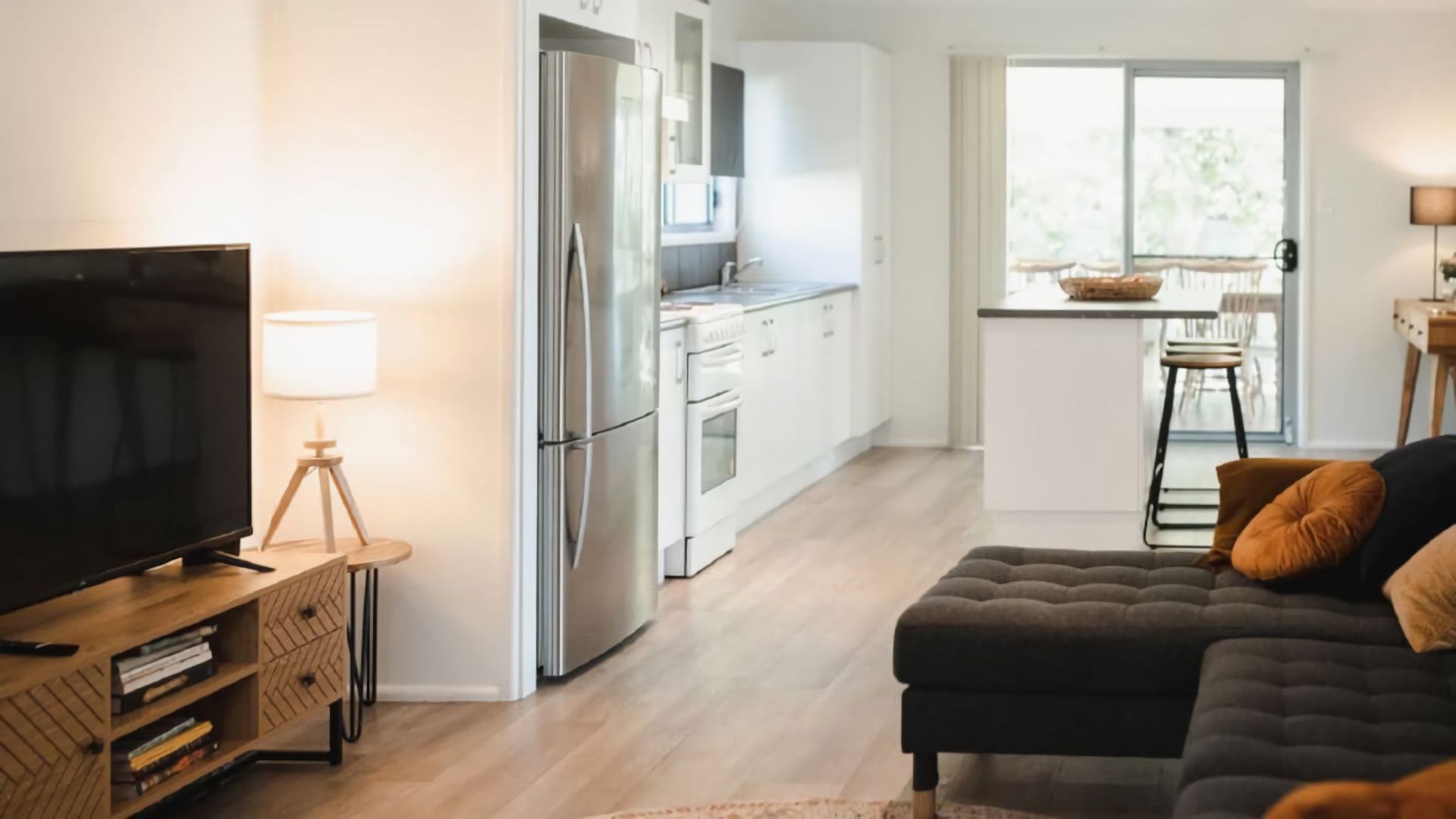 Een moderne woonkamer met een flatscreen televisie, een donkere bank met kussens en een houten bijzettafeltje met een lamp.