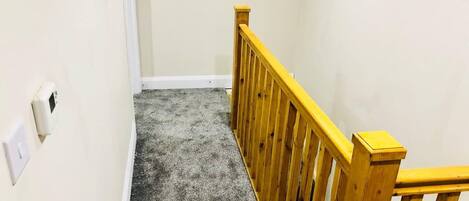 A narrow hallway with a wooden railing, carpeted floor, and a small window at the end.
