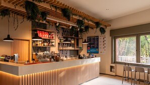 Zona de bar moderna con mostrador de madera, estanterías con botellas y plantas colgantes. Hay dos taburetes de bar y un ventanal con vistas a árboles.
