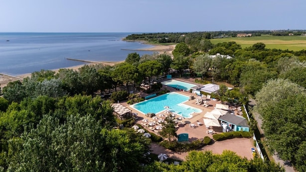Vista aérea de un complejo turístico con una gran piscina rodeada de sillas de descanso y sombrillas, una playa y exuberante vegetación.