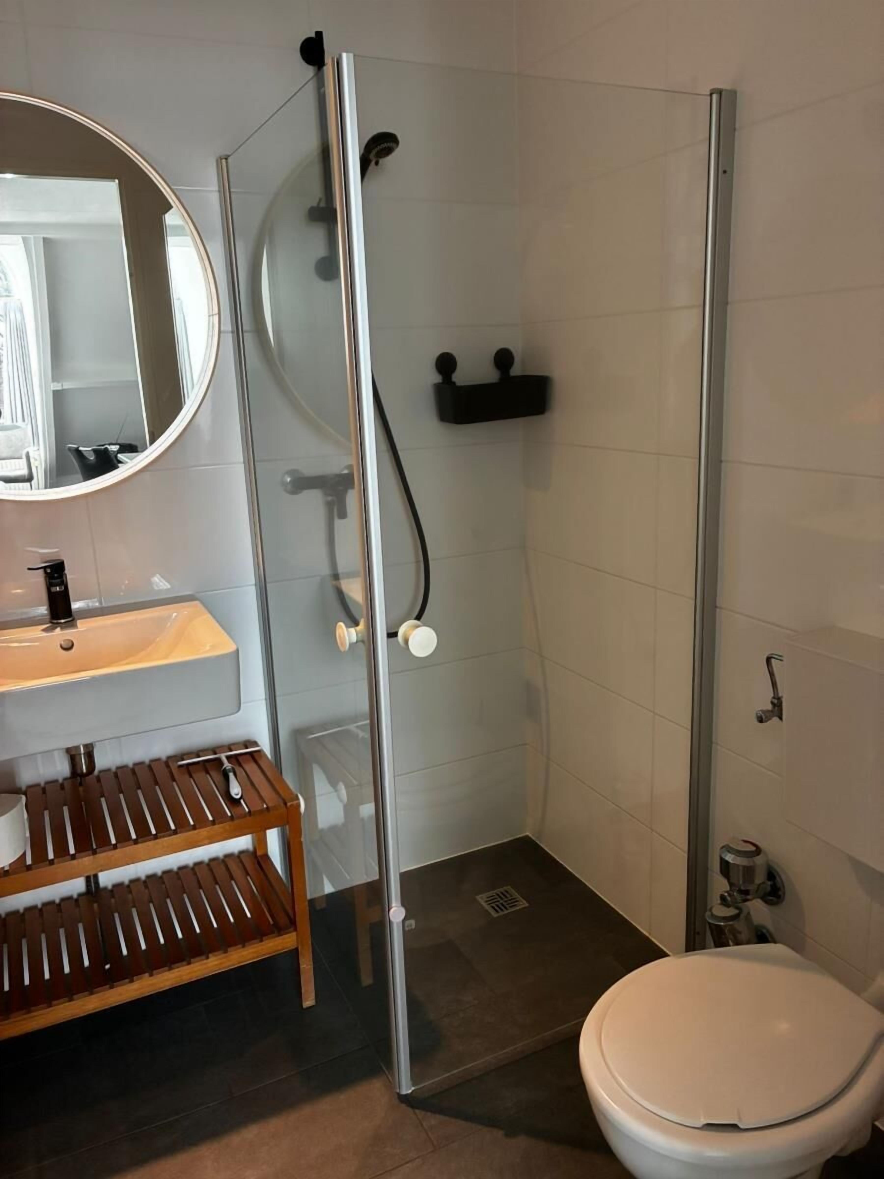 A bathroom with a glass-enclosed shower, a round mirror, a wooden vanity, and a toilet.