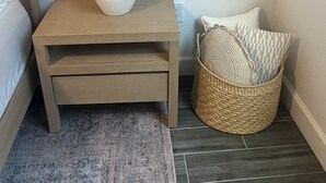 Une chambre d’angle avec une table de chevet en bois, une lampe blanche, un panier en osier contenant des coussins et un tapis à motifs.