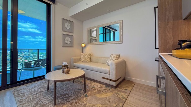 A modern living room with a sofa, a coffee table, and a large window offering a city view. - Luxury View-Modern 1-BR Condo with Panoramic Ocean Views, Pool & Gym–Miami Beach (Miami Beach)