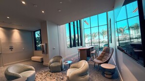 A modern lounge area with comfortable seating, a small table, and large windows offering a view of palm trees and a building. - Luxury View-Modern 1-BR Condo with Panoramic Ocean Views, Pool & Gym–Miami Beach (Miami Beach)
