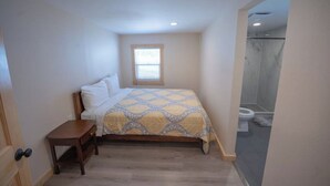 Un dormitorio con cama, mesita de noche, ventana y vista al baño.