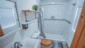 A bathroom with a toilet, sink, shower, and a small shelf with a plant.