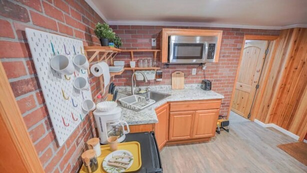A compact kitchen with a brick wall, wooden cabinets, a microwave, and a pegboard with mugs.