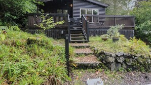 A cabin with a wooden deck, a staircase leading to the entrance, and a stone wall surrounding the property.
