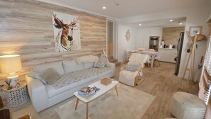 Un salon moderne avec un canapé, une table basse et un tableau représentant un cerf. On y trouve également un coin repas avec une table et des chaises, et une cuisine à l’arrière-plan.