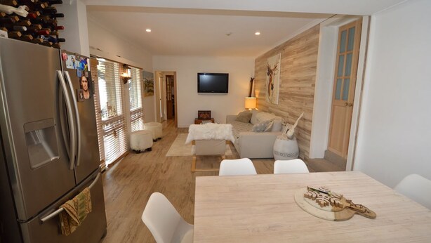 Une cuisine moderne dotée d’un réfrigérateur intégré et d’un meuble à vin, un coin repas avec une table et des chaises en bois, un salon avec un canapé et un téléviseur, et un couloir avec un plancher en bois et une porte donnant sur une autre pièce.