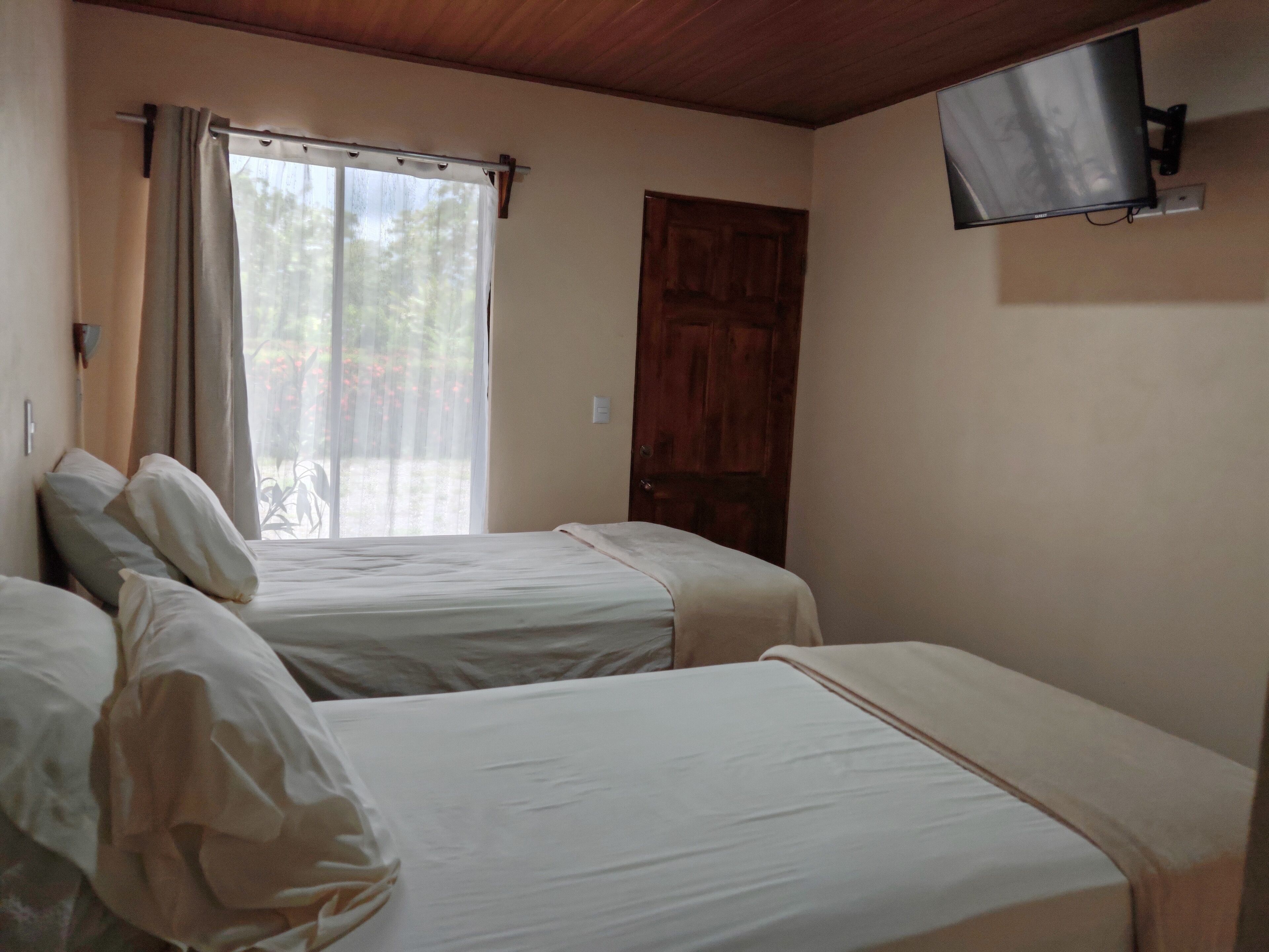 Une chambre avec deux lits simples, une porte en bois, un téléviseur fixé au mur et une fenêtre avec des rideaux.
