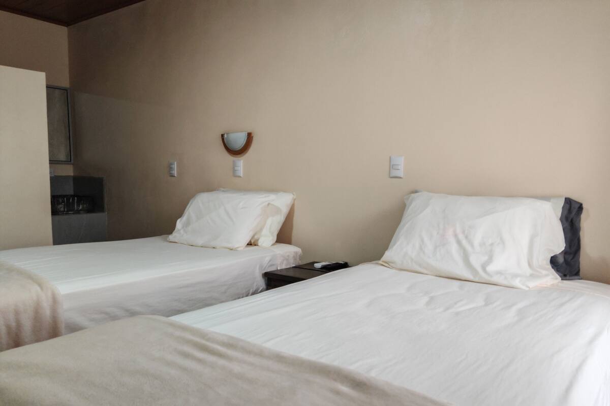 Une chambre d’hôtel avec deux lits simples, une petite table, un luminaire fixé au mur et, au fond, une kitchenette.